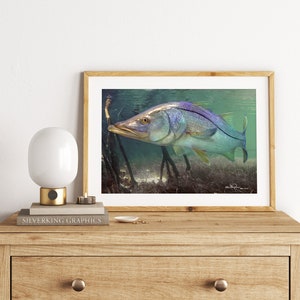 Pode incluir: Uma impressão de arte emoldurada com uma representação subaquática detalhada de um peixe Snook. O peixe tem um corpo prateado com uma faixa roxa e barbatanas amarelas. A obra de arte é definida contra um fundo de água turva e raízes de mangue.