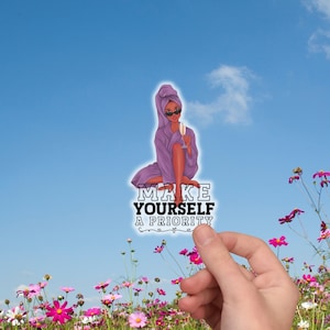 May include: A sticker with the text "MAKE YOURSELF A PRIORITY" featuring an illustration of a person in a purple robe and towel, holding a champagne glass. The sticker is held in front of a field of flowers and a blue sky.