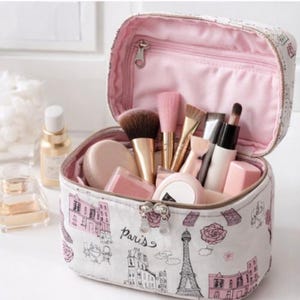 May include: Cosmetic bag with a white exterior and pink lining, decorated with a Paris design including the Eiffel Tower and building sketches. The bag is open, displaying makeup brushes, compacts, and beauty products.