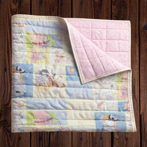 May include: A patchwork baby quilt with a light pink backing and a cream-colored border. The front features squares with animal and tropical designs in pastel colors, including zebras, giraffes, and palm trees.