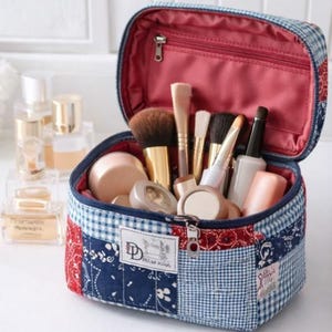May include: Open makeup bag with a patchwork design in red, white, and blue. The bag is filled with various makeup brushes, compacts, and tubes. The interior is lined with red fabric. The bag has a zipper closure and a small label that reads "Dotted Kiss".