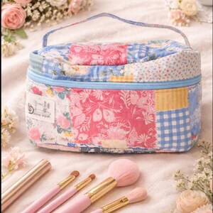 May include: A patchwork cosmetic bag with a blue zipper and handle. The bag features a variety of floral and gingham patterns in pink, blue, and yellow. Makeup brushes with pink handles and gold ferrules are in the foreground.