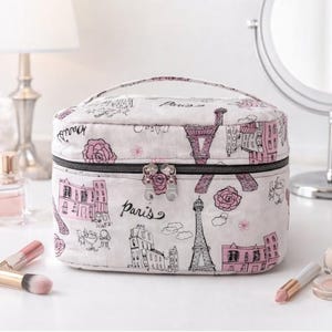 Op de afbeelding: Een witte make-uptas met een zwarte rits en handvat, met een Parijs-thema. De tas is versierd met roze rozen, de Eiffeltoren en Parijse gebouwen. Het woord "Paris" staat op de tas.