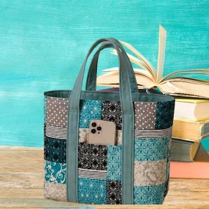 May include: A patchwork tote bag with a teal handle and pocket, holding a smartphone. The bag features a mix of teal, gray, and black patterned fabric. Books are stacked in the background.