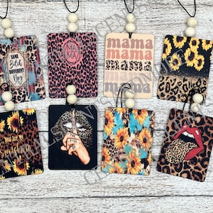 May include: Eight different car air fresheners with various designs. The designs include leopard print, sunflowers, and a woman's lips with a finger over them. The air fresheners are all rectangular and have a black string with wooden beads.