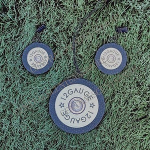May include: A set of three black and gold 12 gauge shotgun shell earrings and necklace. The earrings are small and the necklace is larger. The earrings and necklace are all made of metal and have a black finish with gold accents. The necklace has a black cord.
