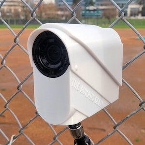 May include: A white security camera with a black lens and the text "THE AVAKIS GLADI" printed on the side. The camera is mounted on a black metal stand and is attached to a chain link fence.