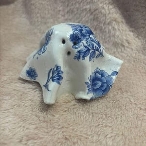 May include: A ceramic ghost figurine with a blue and white floral pattern. The ghost has three small holes for eyes and a whimsical, draped design. The color palette is reminiscent of classic blue and white porcelain.
