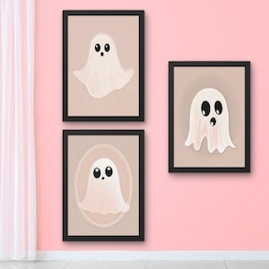 May include: Three framed art prints featuring cartoon ghosts. Each ghost is white with black eyes and a small mouth, set against a beige background. The frames are black, and the prints are displayed on a pink wall.