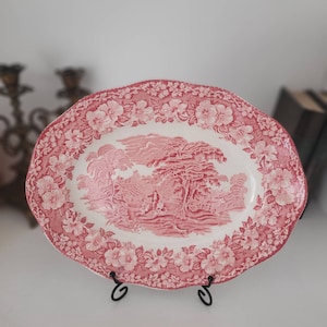 Peut inclure: Un plat de service ovale rose et blanc avec un motif floral et une scène de campagne au centre. Le plat est sur un support noir.