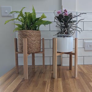 May include: Two wooden plant stands, each supporting a potted plant. One has a woven basket with a green plant, the other a white pot with a flowering plant. The stands are made of light-coloured wood.
