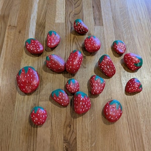 May include: Painted rocks designed to look like strawberries. Each rock is red with white dots and a green leafy top. The rocks are scattered on a wooden surface, creating a playful arrangement.