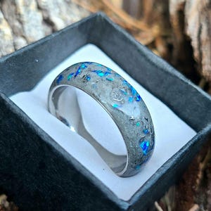 Puede incluir: Un anillo plateado con una banda de material gris incrustada con motas azules y turquesas. El anillo se presenta en una caja negra con interior blanco. El diseño del anillo es moderno y llamativo.
