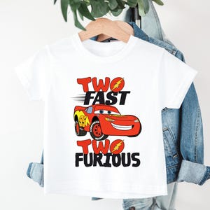 May include: A white short-sleeved t-shirt featuring a graphic of a red race car with the number 95 on its side. The text "TWO FAST" appears above the car and "TWO FURIOUS" appears below, both with yellow lightning bolt accents on the letter O.