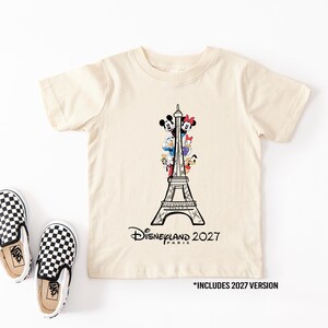 May include: A cream-colored short-sleeve t-shirt featuring a black line drawing of the Eiffel Tower with Disney characters Mickey, Minnie, Donald, Daisy, and Pluto. The text reads Disneyland Paris 2027. Two black and white checkered slip-on shoes.