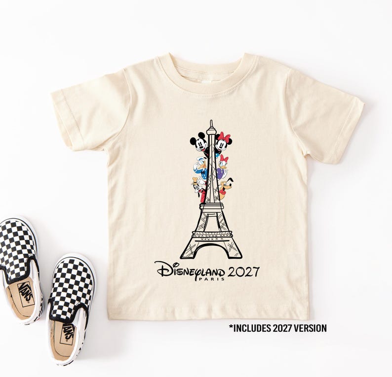 May include: A cream-colored short-sleeve t-shirt featuring a black line drawing of the Eiffel Tower with Disney characters Mickey, Minnie, Donald, Daisy, and Pluto. The text reads Disneyland Paris 2027. Two black and white checkered slip-on shoes.