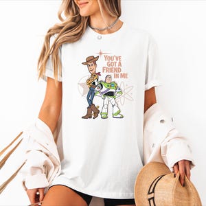May include: A white short-sleeved t-shirt featuring a graphic print of Woody and Buzz Lightyear from Toy Story. The text "YOU'VE GOT A FRIEND IN ME" is printed in a light brown colour above the characters, accompanied by star and atom graphics.