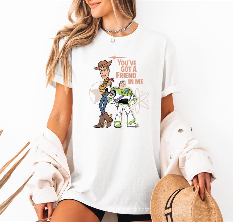 May include: A white short-sleeved t-shirt featuring a graphic print of Woody and Buzz Lightyear from Toy Story. The text "YOU'VE GOT A FRIEND IN ME" is printed in a light brown colour above the characters, accompanied by star and atom graphics.