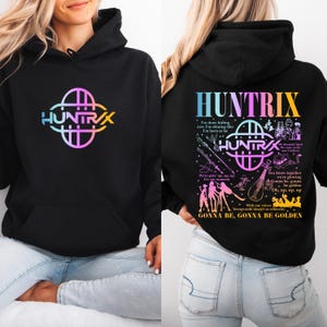 Peut inclure: Sweat à capuche noir avec un design avant et arrière. L'avant présente le mot "HUNTRIX" dans une police dégradée colorée avec un graphique assorti. Le dos affiche le même texte et graphique, ainsi que des paroles de chansons et des illustrations.