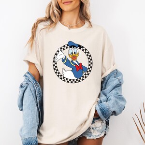Pu&ograve; includere: Una t-shirt beige a maniche corte con una grafica di Paperino all'interno di un cerchio a scacchi bianchi e neri. Il personaggio indossa una camicia da marinaio blu, un papillon rosso e un cappello blu, sorridendo e gesticolando.