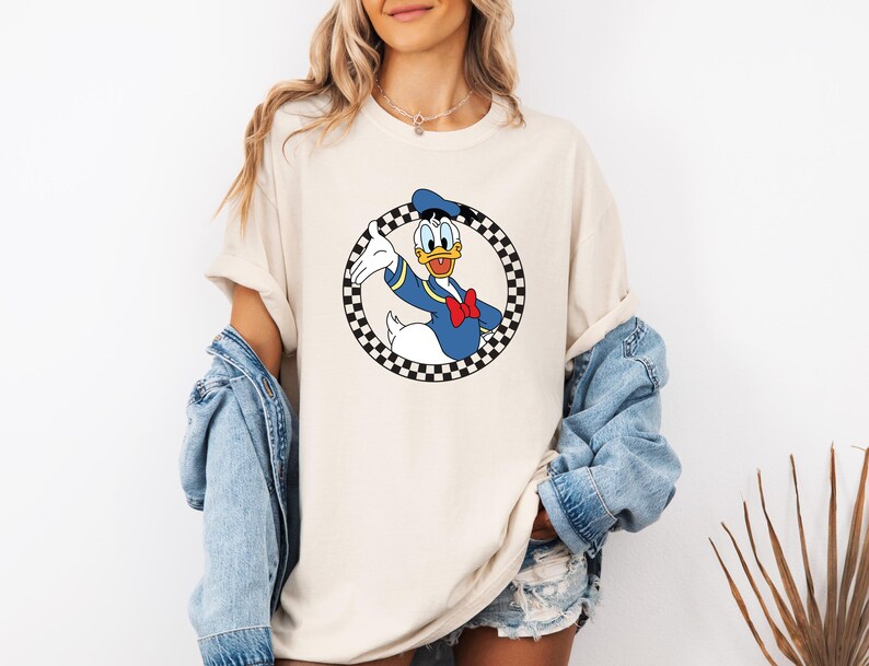 Pu&ograve; includere: Una t-shirt beige a maniche corte con una grafica di Paperino all'interno di un cerchio a scacchi bianchi e neri. Il personaggio indossa una camicia da marinaio blu, un papillon rosso e un cappello blu, sorridendo e gesticolando.
