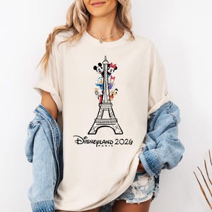 May include: A beige short-sleeve t-shirt featuring a black line-art illustration of the Eiffel Tower with Mickey Mouse, Minnie Mouse, Donald Duck, and Pluto characters. The text Disneyland Paris 2026 is printed in black below the tower graphic.