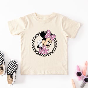 Pu&ograve; includere: Una t-shirt a maniche corte color crema con la grafica di un topo dei cartoni animati con un fiocco e un vestito a pois rosa, racchiusa in un bordo circolare a scacchi bianchi e neri, accompagnata da un paio di scarpe slip-on a scacchi.