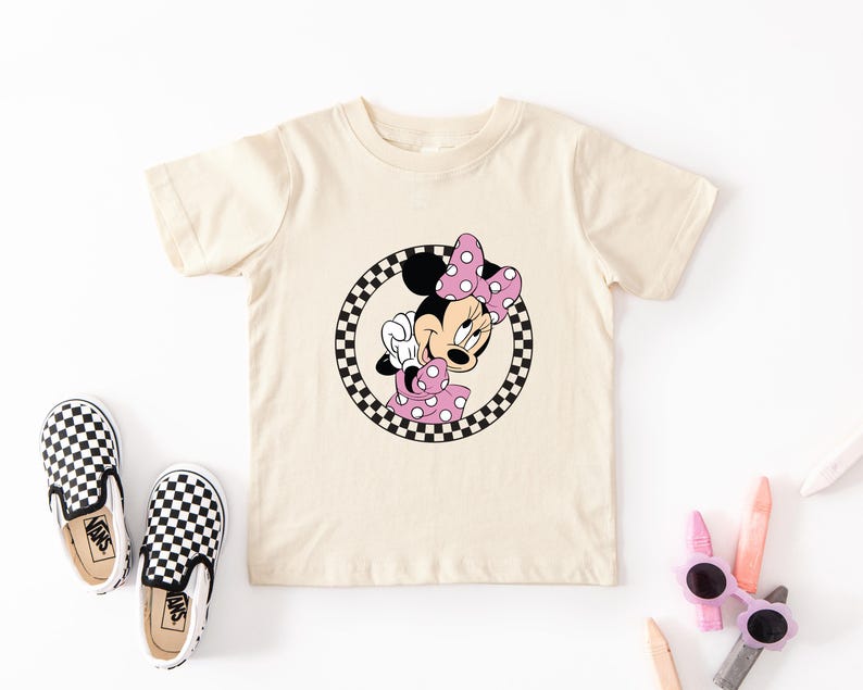 Pu&ograve; includere: Una t-shirt a maniche corte color crema con la grafica di un topo dei cartoni animati con un fiocco e un vestito a pois rosa, racchiusa in un bordo circolare a scacchi bianchi e neri, accompagnata da un paio di scarpe slip-on a scacchi.