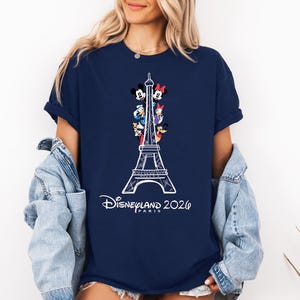 May include: A navy blue short-sleeve t-shirt featuring a white line-art graphic of the Eiffel Tower with various cartoon characters positioned within the structure. Below the tower, the text reads Disneyland 2026 PARIS in a white script font.