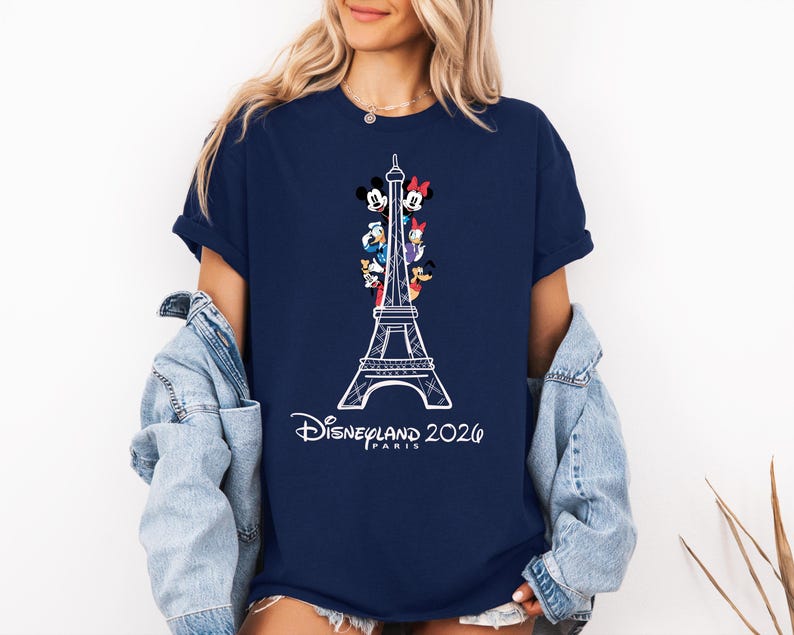 May include: A navy blue short-sleeve t-shirt featuring a white line-art graphic of the Eiffel Tower with various cartoon characters positioned within the structure. Below the tower, the text reads Disneyland 2026 PARIS in a white script font.