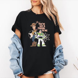 May include: A black short-sleeved t-shirt featuring a graphic print of Woody and Buzz Lightyear standing side by side. The text "YOU'VE GOT A FRIEND IN ME" is printed in a light peach colour above the characters, accompanied by star and atom icons.