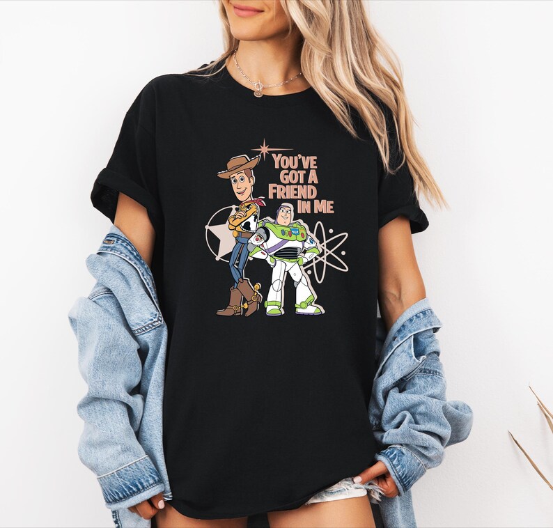 May include: A black short-sleeved t-shirt featuring a graphic print of Woody and Buzz Lightyear standing side by side. The text "YOU'VE GOT A FRIEND IN ME" is printed in a light peach colour above the characters, accompanied by star and atom icons.