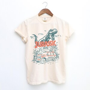 Pu&ograve; includere: Una t-shirt a maniche corte color crema con una grafica verde scuro di un Tyrannosaurus Rex ruggente sopra un veicolo di Jurassic Park e palme. Il testo JURASSIC PARK &egrave; stampato in arancione e la parola JURASSIC appare sul veicolo.