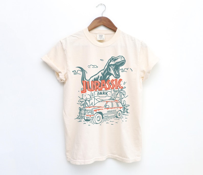 Pu&ograve; includere: Una t-shirt a maniche corte color crema con una grafica verde scuro di un Tyrannosaurus Rex ruggente sopra un veicolo di Jurassic Park e palme. Il testo JURASSIC PARK &egrave; stampato in arancione e la parola JURASSIC appare sul veicolo.