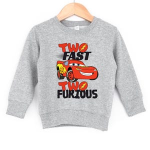 May include: A heather gray long-sleeve sweatshirt featuring a graphic of a red race car with the number 95 on the side. The text "TWO FAST" appears above the car and "TWO FURIOUS" appears below, with lightning bolt icons in the letter O.
