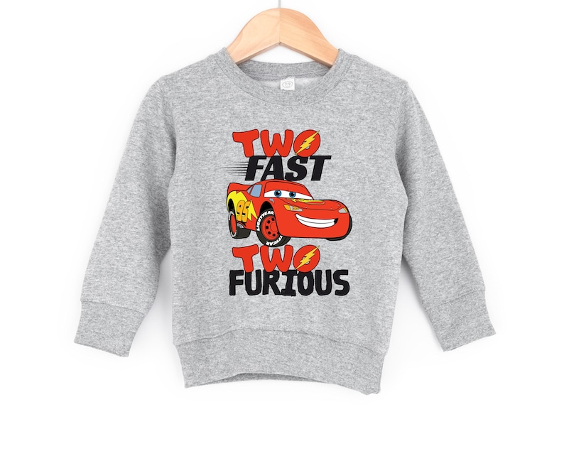 May include: A heather gray long-sleeve sweatshirt featuring a graphic of a red race car with the number 95 on the side. The text "TWO FAST" appears above the car and "TWO FURIOUS" appears below, with lightning bolt icons in the letter O.