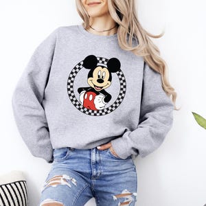 Pu&ograve; includere: Una felpa girocollo grigio chiaro con una grafica di Topolino centrata all'interno di un cerchio a scacchi bianco e nero. Il personaggio &egrave; raffigurato con le sue tipiche orecchie nere, pantaloncini rossi e guanti bianchi sul tessuto grigio.