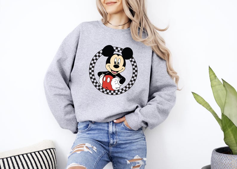 Pu&ograve; includere: Una felpa girocollo grigio chiaro con una grafica di Topolino centrata all'interno di un cerchio a scacchi bianco e nero. Il personaggio &egrave; raffigurato con le sue tipiche orecchie nere, pantaloncini rossi e guanti bianchi sul tessuto grigio.