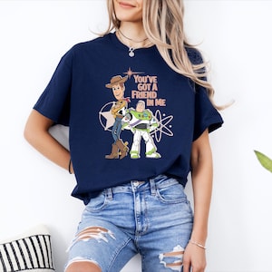 May include: A navy blue short-sleeve t-shirt featuring a graphic print of Woody and Buzz Lightyear from Toy Story. The text "YOU'VE GOT A FRIEND IN ME" is printed in light peach above the characters, accompanied by white star and orbit graphics.