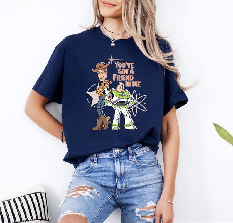 May include: A navy blue short-sleeve t-shirt featuring a graphic print of Woody and Buzz Lightyear from Toy Story. The text "YOU'VE GOT A FRIEND IN ME" is printed in light peach above the characters, accompanied by white star and orbit graphics.
