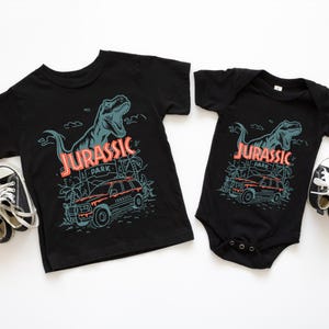 Pu&ograve; includere: Una t-shirt nera a maniche corte e un body nero a maniche corte, entrambi con una stampa grafica di un Tyrannosaurus Rex, una scena nella giungla e un veicolo con il testo JURASSIC PARK. Due paia di scarpe da ginnastica bianche e nere sono vicine.