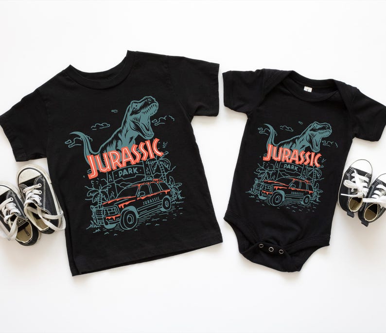 Pu&ograve; includere: Una t-shirt nera a maniche corte e un body nero a maniche corte, entrambi con una stampa grafica di un Tyrannosaurus Rex, una scena nella giungla e un veicolo con il testo JURASSIC PARK. Due paia di scarpe da ginnastica bianche e nere sono vicine.
