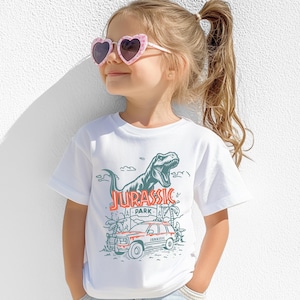 Pu&ograve; includere: Una t-shirt bianca a maniche corte con una stampa grafica di un Tyrannosaurus Rex, un veicolo e il testo Jurassic Park in inchiostro rosso e verde scuro. Il design include palme e nuvole, centrato sulla parte anteriore del capo.