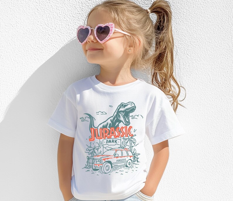 Pu&ograve; includere: Una t-shirt bianca a maniche corte con una stampa grafica di un Tyrannosaurus Rex, un veicolo e il testo Jurassic Park in inchiostro rosso e verde scuro. Il design include palme e nuvole, centrato sulla parte anteriore del capo.