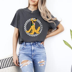 Pu&ograve; includere: Una t-shirt a maniche corte grigio antracite con una grafica del cane dei cartoni animati Pluto in giallo con un collare verde, centrata all'interno di un bordo circolare a scacchi bianchi e neri. La t-shirt ha un girocollo e una vestibilit&agrave; ampia.