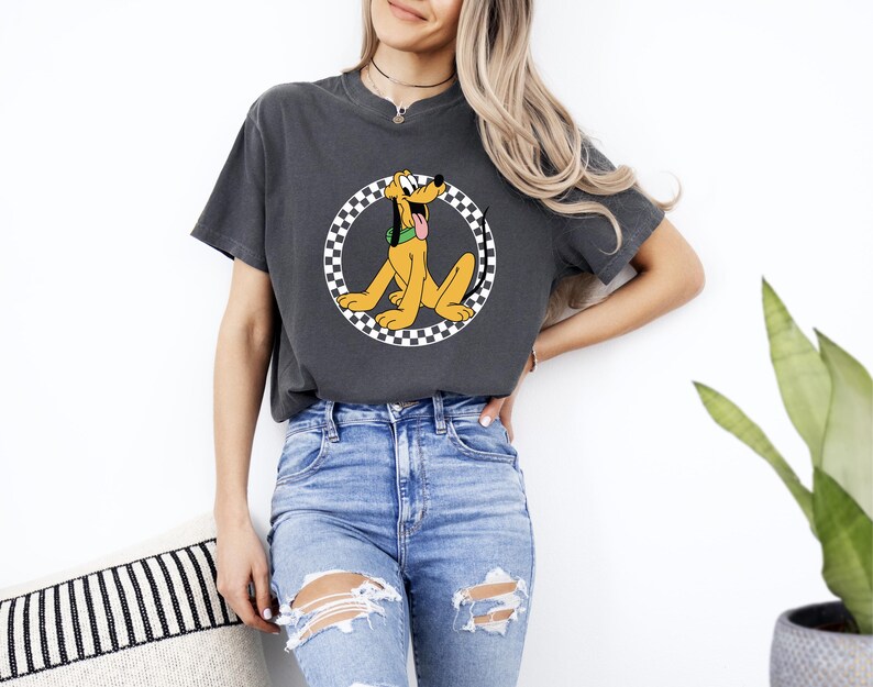 Pu&ograve; includere: Una t-shirt a maniche corte grigio antracite con una grafica del cane dei cartoni animati Pluto in giallo con un collare verde, centrata all'interno di un bordo circolare a scacchi bianchi e neri. La t-shirt ha un girocollo e una vestibilit&agrave; ampia.