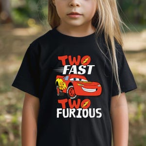 May include: A black short-sleeved t-shirt featuring a graphic of a red race car with the number 95 on the side. The text "TWO FAST" appears above the car, and "TWO FURIOUS" appears below, both incorporating yellow lightning bolt icons.