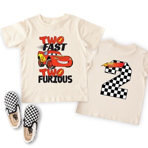 May include: Two cream-colored short-sleeve t-shirts and a pair of black and white checkered slip-on shoes. One shirt features a red race car graphic with the text TWO FAST TWO FURIOUS. The other shirt displays a large checkered number 2.
