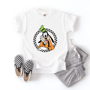 Pu&ograve; includere: Una t-shirt bianca a maniche corte con una grafica del personaggio dei cartoni animati Pippo all'interno di un cerchio a scacchi bianchi e neri, abbinata a pantaloni della tuta grigi e un paio di scarpe slip-on a scacchi con suola bianca.