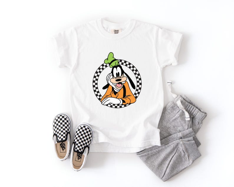 Pu&ograve; includere: Una t-shirt bianca a maniche corte con una grafica del personaggio dei cartoni animati Pippo all'interno di un cerchio a scacchi bianchi e neri, abbinata a pantaloni della tuta grigi e un paio di scarpe slip-on a scacchi con suola bianca.